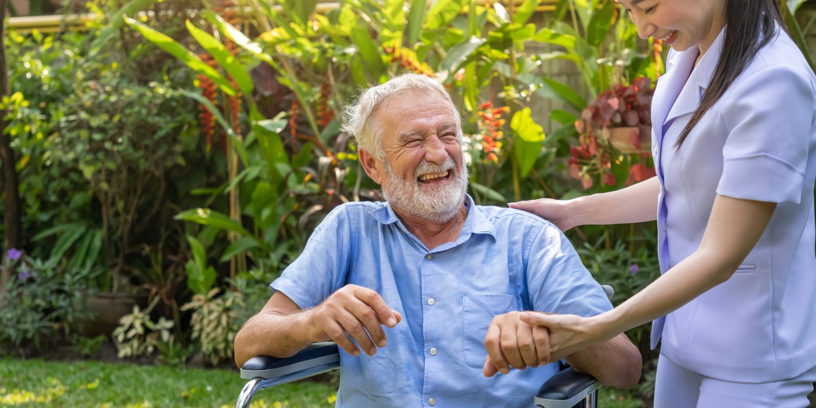 Happy nurse holding laughing elderly man hand on wheelchair in g